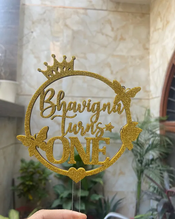 This glittery gold topper for "Bhavigna Turns ONE" is all about sparkle and celebration. The design incorporates a crown and butterflies for a truly royal and whimsical first birthday theme.