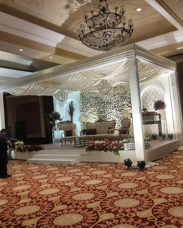 A luxurious indoor wedding stage featuring a white mandap structure against a full wall of white flowers. The setup is completed with traditional seating and enhanced by the hall's ornate crystal chandelier.