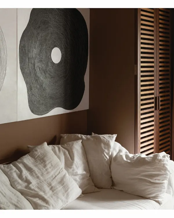 A comfortable slipcover sofa sits against a warm, earthy brown wall, creating a cozy corner for relaxation. The large, abstract artwork and slatted wooden screen add layers of texture and interest, defining the room's calm and artistic character.