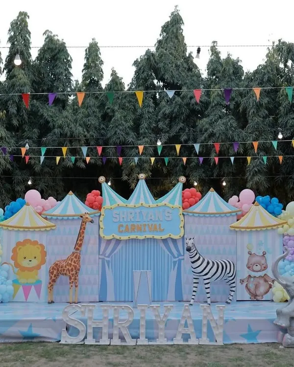 The main stage for a pastel carnival themed first birthday. This outdoor setup features a backdrop designed like circus tents with cute animal characters.