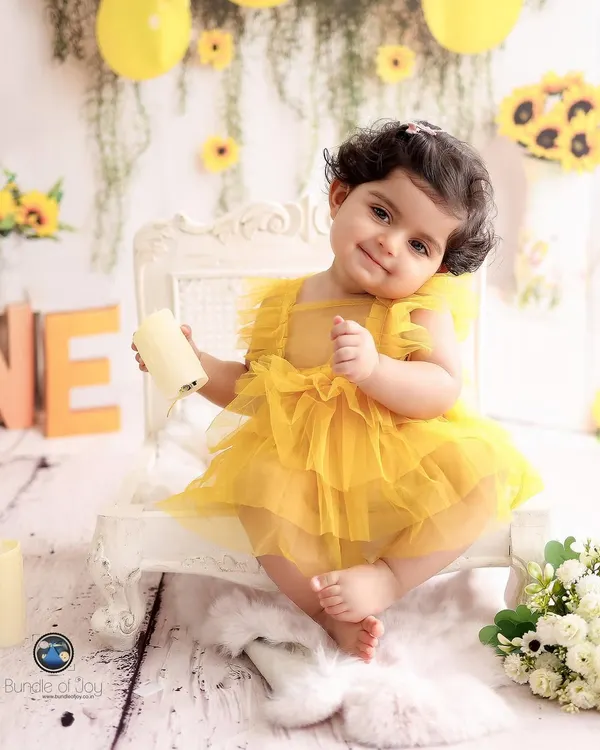 A charming portrait of a one-year-old girl in a bright yellow dress, sitting on a miniature vintage chair as part of her sunflower themed birthday shoot.