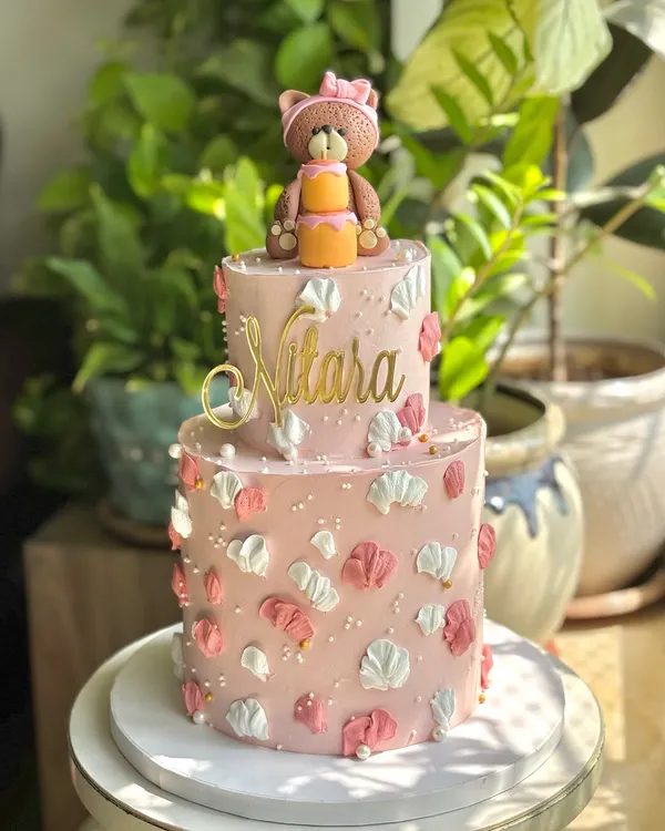 A little pink dream for a sweet one-year milestone. This two-tier cake features palette-knife florals, a custom gold name topper, and an adorable, hand-crafted teddy bear holding her own mini cake.