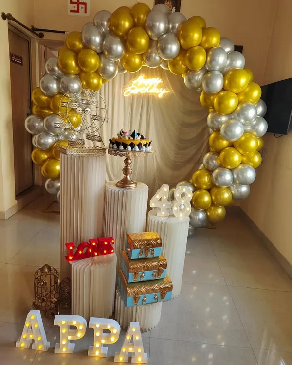 For this 48th birthday, I created a classy gold and silver balloon arch to frame the celebration space. The setup features custom marquee letters spelling "APPA", light-up numbers for the age, and a "Happy Birthday" neon sign, all brought together with decorative pedestals and props.