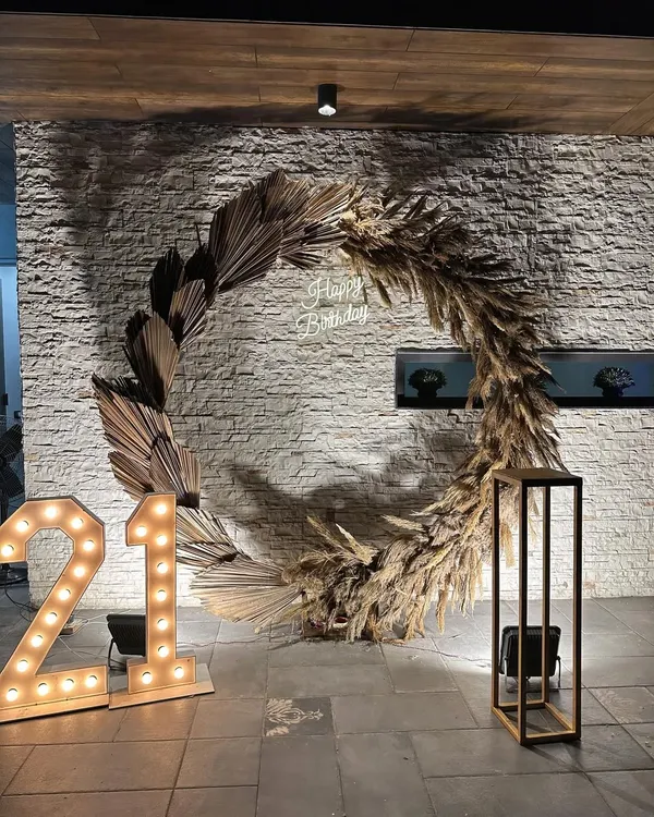 A chic and rustic setup for a 21st birthday party, featuring a large pampas grass wreath, a neon sign, and marquee lights.
