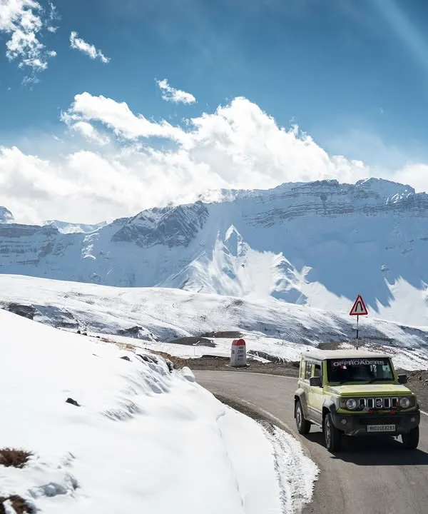 A bright yellow Suzuki Jimny navigates a winding road through a vast, snow-covered mountain landscape in Spiti Valley. This shot captures the essence of a true 4x4 adventure photoshoot, placing the vehicle in its element.
