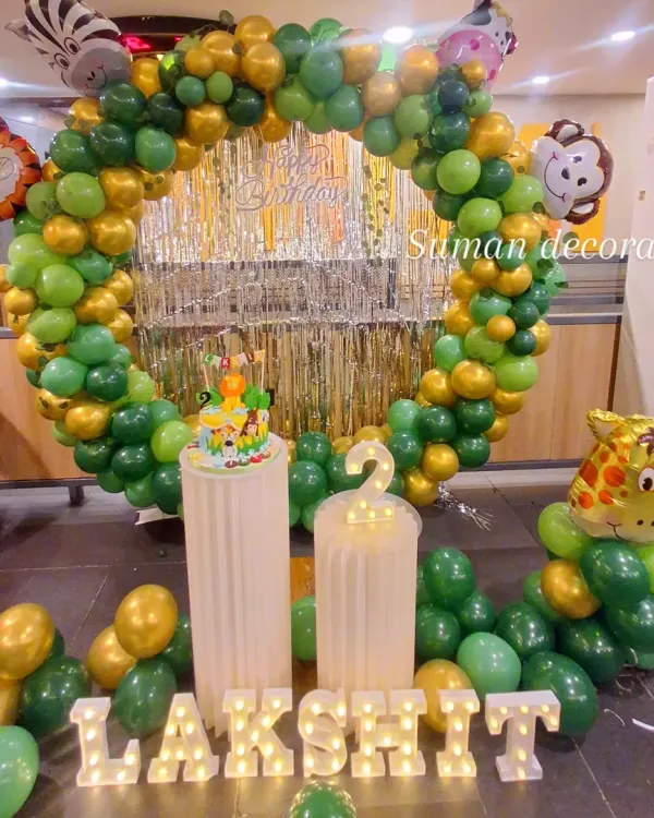A fun jungle safari theme for a second birthday. This setup features a circular balloon arch in shades of green and gold, a silver fringe backdrop, animal character cutouts like a monkey and giraffe, and light-up marquee letters.