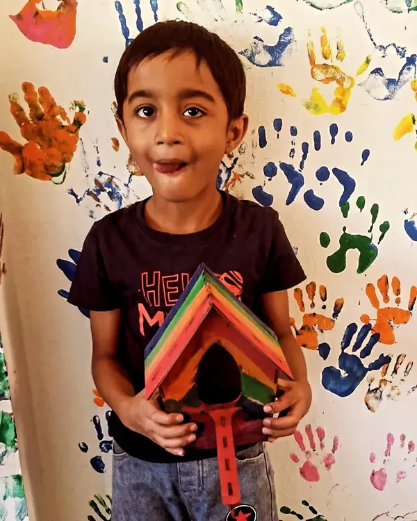 A proud young builder shows off the rainbow-colored birdhouse he made, standing in front of our signature handprint wall.