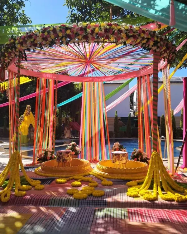 A poolside Haldi setup in Jim Corbett, combining colorful ribbon drapes with traditional marigold garlands. The low seating (patlas) and floral arch create a perfect space for the ceremony.