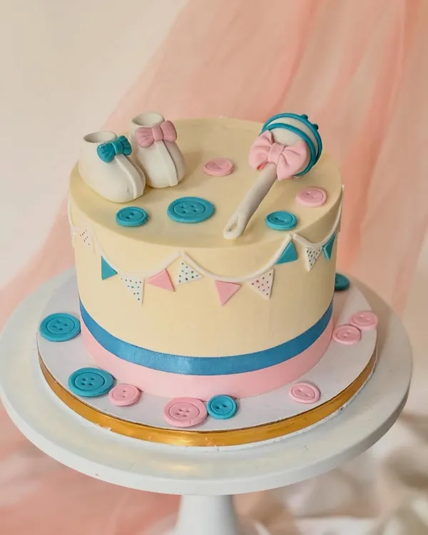 A classic baby shower cake with a gender reveal theme. This cake is decorated with pink and blue buttons, a bunting banner, and adorable baby booties and a rattle on top.