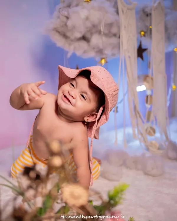 An adorable and playful shot of a baby with a huge smile, wearing a cute pink hat. The dreamy background with clouds and stars adds a whimsical touch to this indoor baby shoot.