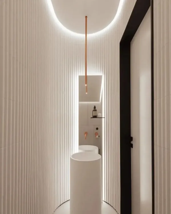 A minimalist powder room design featuring curved, fluted walls and a sleek pedestal sink. The vertical pendant light and backlighting create a soft, ambient glow, making the small space feel special.