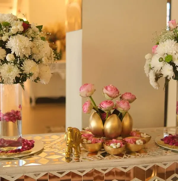 An elegant welcome table setup featuring tall glass vases with large bouquets of white chrysanthemums. The centerpiece includes pink lotus buds in golden egg-shaped vases.
