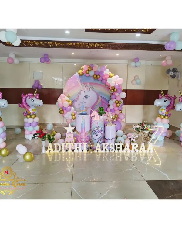 A spectacular unicorn-themed party setup with a large unicorn backdrop, pastel balloon arch, and unicorn-shaped balloon columns. The floor is decorated with scattered balloons and marquee letters for the names.