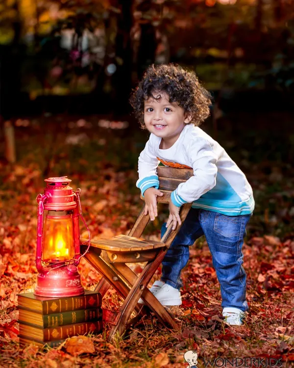 A toddler boy with amazing curly hair leans on a chair in a forest setting, surrounded by autumn leaves and a glowing lantern. The warm, magical light creates a stunning outdoor portrait.