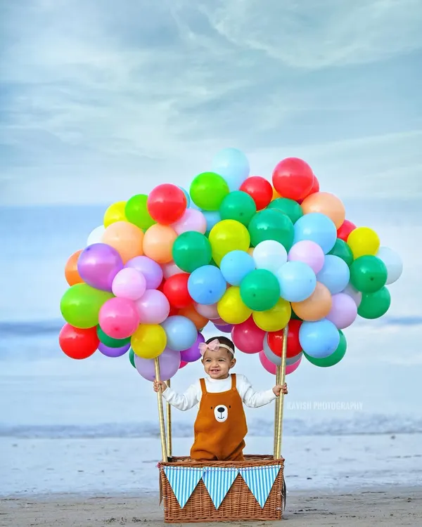 A creative beach photoshoot featuring a happy baby girl in a whimsical hot air balloon basket made of balloons. This is an example of a fun, themed milestone session.