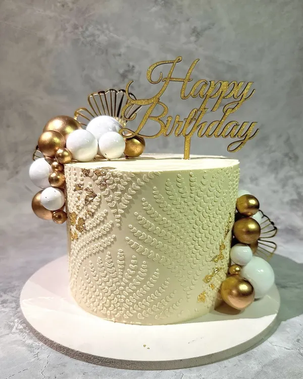An example of detailed buttercream work. This cake features an intricate white-on-white stenciled pattern, accented with clusters of gold and white spheres for a sophisticated finish.