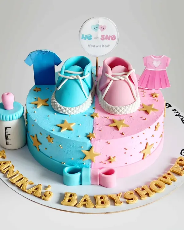A "He or She" gender reveal cake designed as a heart. One side is blue for a boy and the other is pink for a girl, topped with adorable baby shoes and clothes.