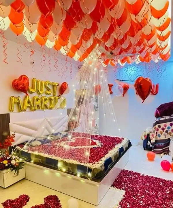 A "Just Married" first night room decoration. The ceiling is covered with red and white balloons, the bed is decorated with a heart made of rose petals, and foil balloons spell out the happy message.