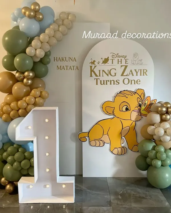 A 'Hakuna Matata' first birthday featuring a Lion King theme. This setup includes a large light-up number '1', a custom arched backdrop with Simba, and a balloon garland in safari-inspired colors like olive green and tan.