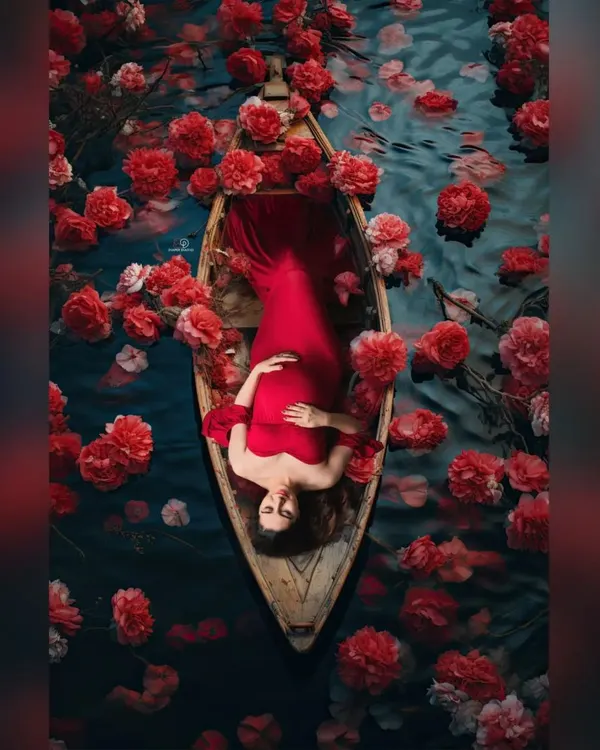A breathtaking overhead shot of a mom-to-be in a red gown, floating in a boat filled with matching red flowers. This is a highly artistic and memorable concept for a maternity shoot.