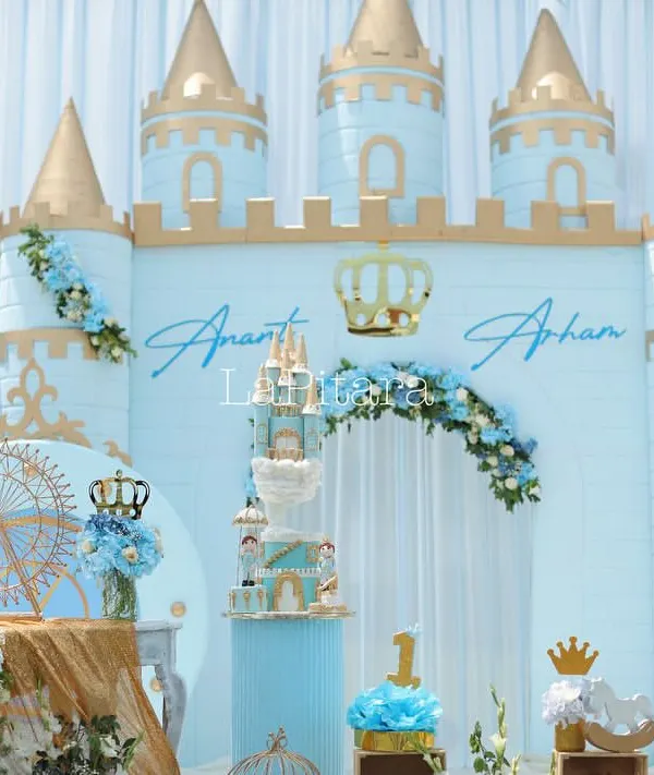 The main stage of a royal prince-themed birthday, featuring a magnificent castle backdrop in pastel blue and gold. The setup includes a tiered cake display, personalized names, and floral arrangements, creating a picture-perfect focal point for the celebration.