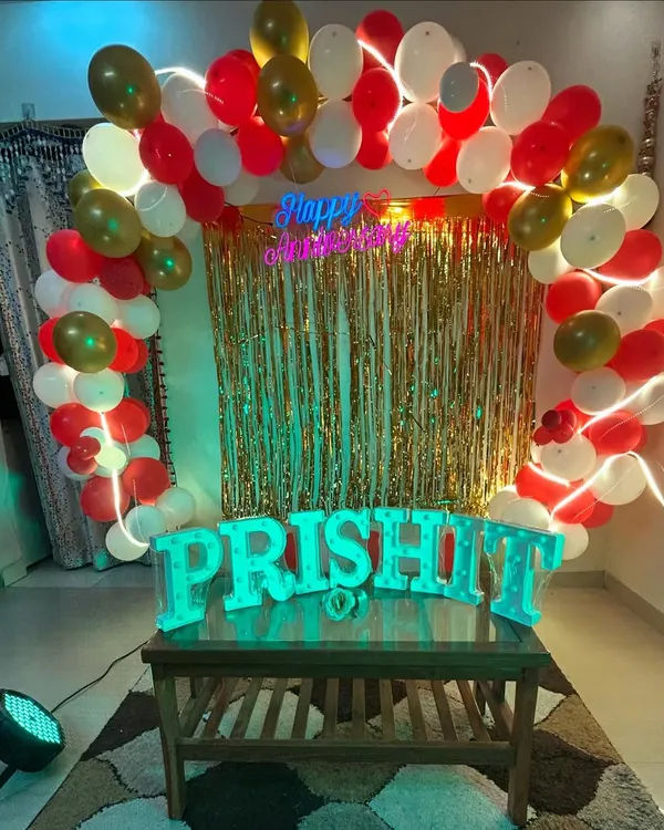 A beautiful anniversary decoration setup for a home party. It features a balloon arch in red, white, and gold, a shimmer curtain backdrop, a neon sign, and light up letters for the couple's name.