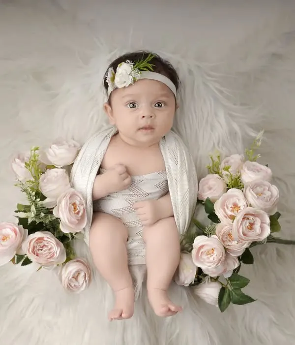 A beautiful, wide-eyed baby girl looking right at the camera. She is wrapped in white and surrounded by soft pink roses, creating a sweet and angelic portrait.
