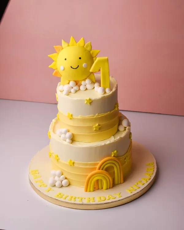 A burst of sunshine for a first birthday celebration. This cheerful two-tier cake features a bright yellow and white design with fondant suns, clouds, and rainbows, perfect for celebrating your little one's big day.