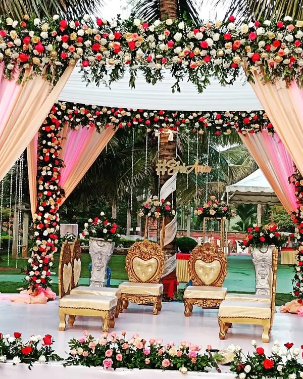 A beautiful outdoor mandap setup with pink and peach drapes, surrounded by lush greenery and palm trees.