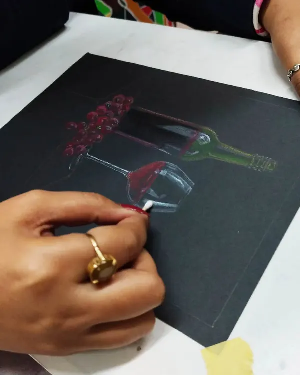 A student creating a still life of a wine bottle and glass using oil pastels on a black sheet. This technique teaches how to render light and reflection on dark surfaces.