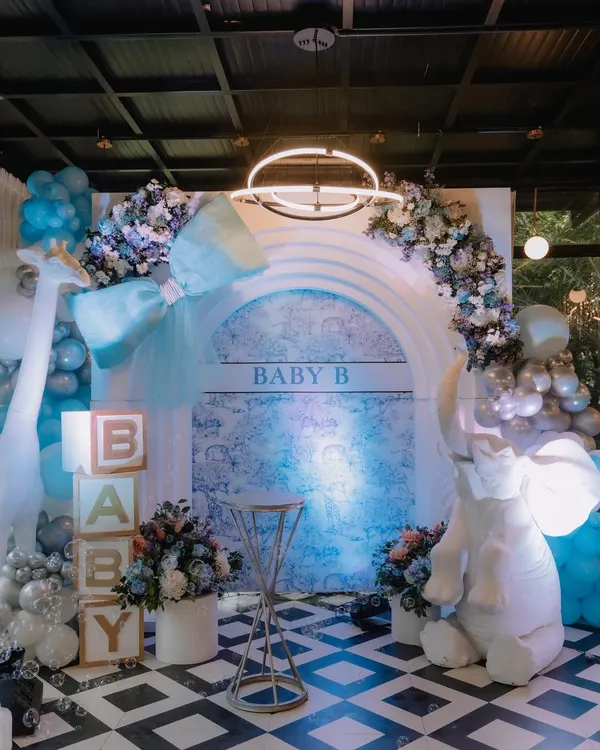 A stunning Dior-inspired baby shower stage in cool shades of blue, white, and silver. The setup includes a large elephant prop, 'BABY' blocks, and a beautiful floral and balloon arch over a custom backdrop.