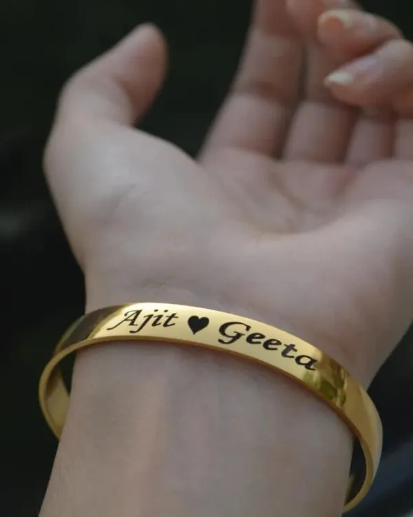 A close-up of our gold-plated cuff bracelet, engraved with the names 'Ajit' and 'Geeta' separated by a heart symbol. This is one of my most requested styles for couples celebrating their bond.