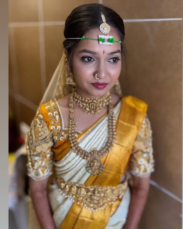 A classic South Indian bridal look, featuring a sharp wing liner and a bold lip that perfectly matches the bride's white and gold Kanjivaram saree. The look is completed with traditional temple jewelry and a matha patti.