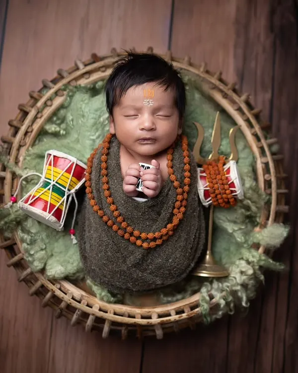 A classic 'Har Har Mahadev' shot. The baby is safely swaddled and posed in a basket, surrounded by symbolic items like the trishul and damaru, creating a serene and powerful image.