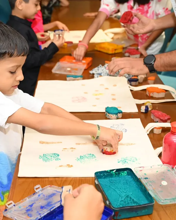 A close-up view of children's hands using colorful stamps to decorate tote bags. We use child-safe fabric paints and a variety of fun tools to make the experience tactile and exciting for young artists.