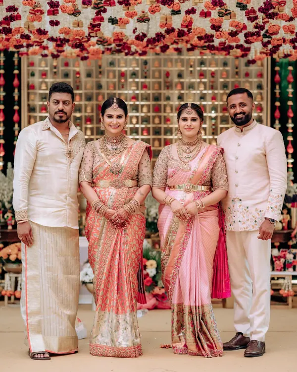 A beautiful family portrait from a naming ceremony. The sisters are coordinated in our bespoke pink and peach Kanchipuram silk sarees with heavily embroidered blouses, creating a harmonious and elegant look.