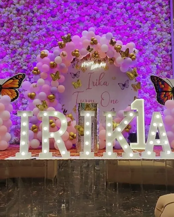 A vibrant butterfly-themed first birthday stage. This setup features a custom backdrop, a full organic balloon arch in pink and purple, and large light-up marquee letters spelling out the birthday girl's name, creating a magical photo opportunity.