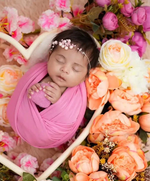 A newborn rests peacefully inside a heart-shaped basket, surrounded by a beautiful arrangement of pink and cream peonies. This classic newborn photography pose highlights the delicate and precious nature of your baby's first days.