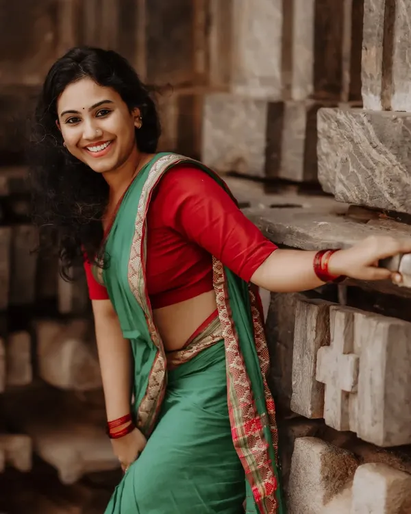 A radiant portrait of a woman in a beautiful green and red saree. Her bright, genuine smile is captured against a rustic stone wall, showcasing a natural and joyful moment.