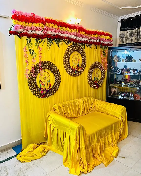 This is a popular setup for a home mehndi or haldi ceremony. It features a bright yellow fabric backdrop, a matching yellow cover for the sofa, and decorative rings to add a modern touch.