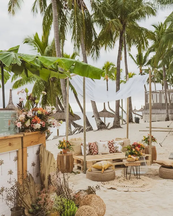 A relaxed beachside lounge in Mauritius, with a canopy made of banana leaves and seating made from wooden pallets and jute poufs.