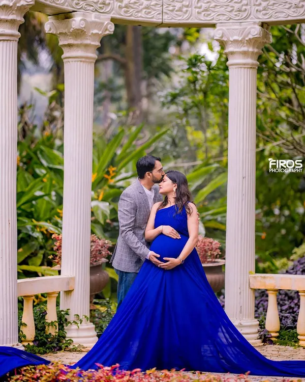 A tender moment between parents-to-be, framed by classic white pillars in a garden setting. The royal blue gown adds a pop of color, highlighting the beautiful baby bump in this serene maternity photograph.