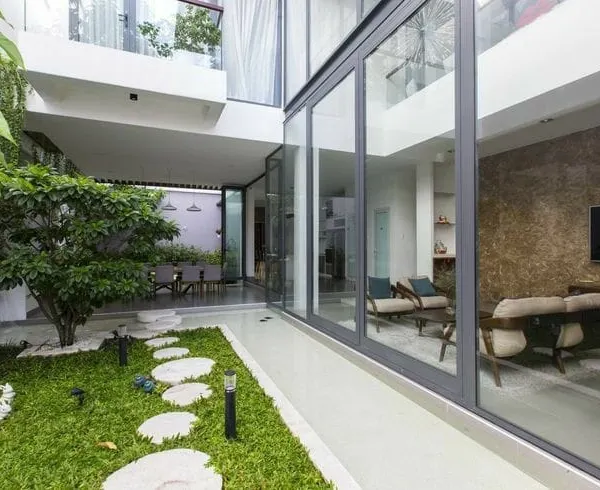 This is a semi-indoor garden that connects the home's interior with the outdoors. During the day, it's a bright, sunlit space with a natural lawn and stone pathway, viewed through large glass walls.