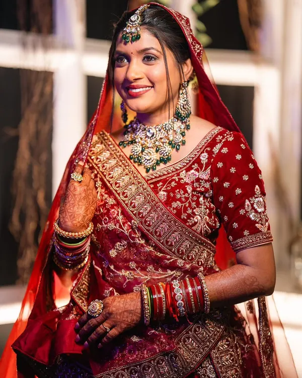 A radiant North Indian bride in a traditional red velvet lehenga. The airbrush makeup provides a flawless base that complements the heavy embroidery and jewelry.
