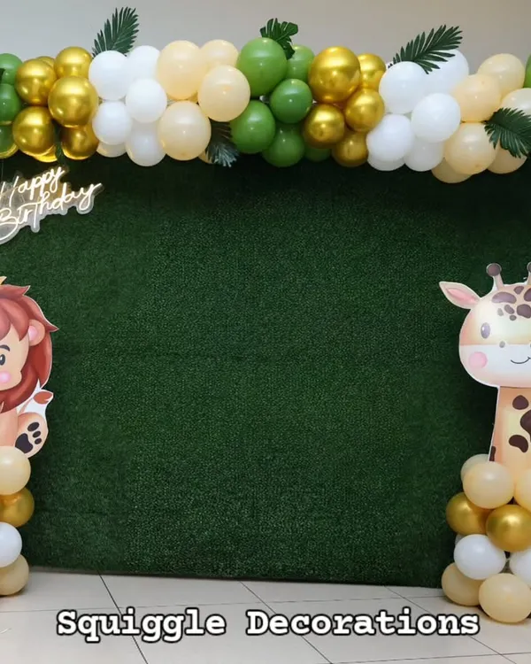 A fun jungle-themed photo booth with an artificial grass backdrop, a 'Happy Birthday' sign, and cute lion and giraffe cutouts. The green, gold, and white balloon garland adds a touch of celebration.