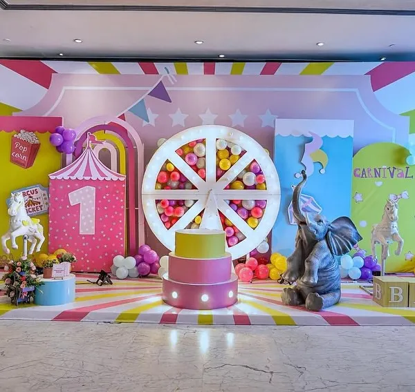 This vibrant carnival stage is the heart of the celebration, designed to be the perfect photo backdrop. It features a custom-lit ferris wheel filled with balloons, a three-tiered cake stand, and life-sized props like a cheerful elephant, creating a truly immersive experience for a first birthday party.