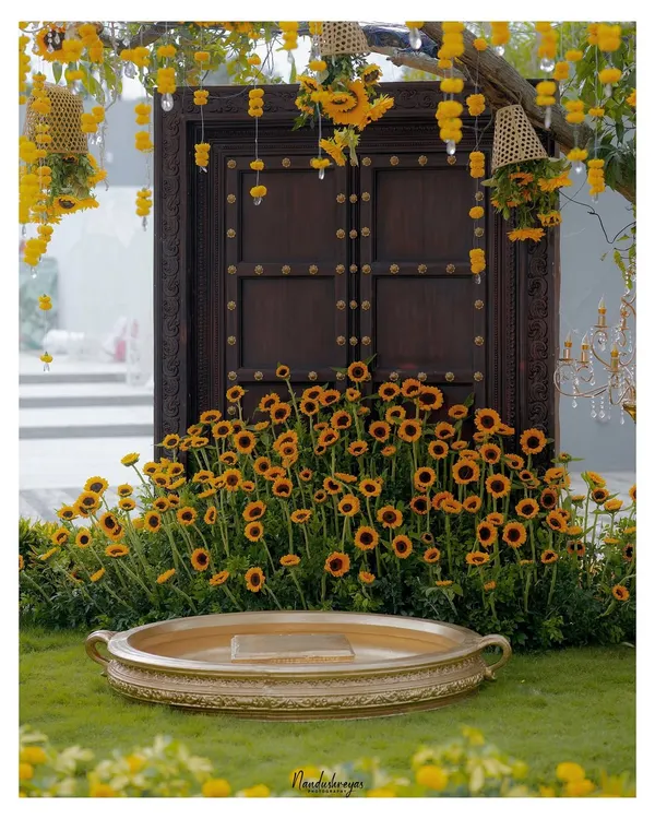 The stunning Haldi ceremony setup, featuring a traditional wooden door, a field of sunflowers, and a brass vessel, all ready for the celebration.