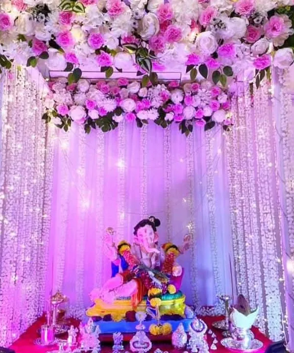 A beautiful home mandap created for the Ganpati festival, featuring a full backdrop of pink and white artificial flowers. The setup is illuminated with warm fairy lights and cascading strings of white floral laris, creating a divine and welcoming space for Bappa.