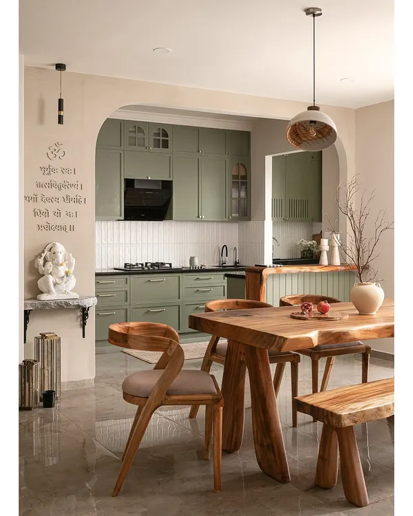 This kitchen features olive green shaker-style cabinets and a live-edge breakfast counter that brings organic charm. The entrance is framed by an elegant arch with limewash walls, and a minimal puja unit sits serenely to one side.