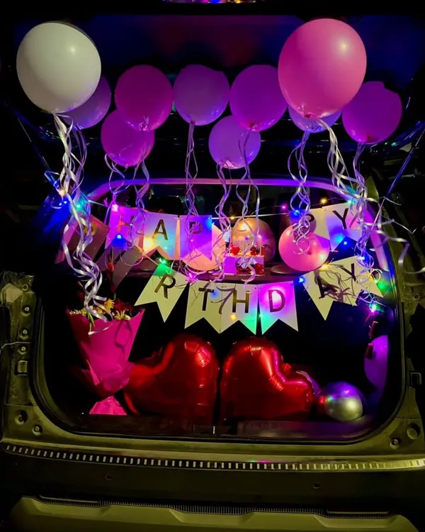 A full view of a birthday car trunk surprise. The back of the car is filled with pink and white balloons, a "Happy Birthday" banner lit by fairy lights, and two large red heart balloons.
